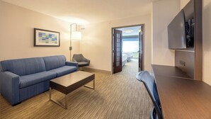 Une chambre d’hôtel avec un canapé bleu, une table basse en bois, une lampe, un téléviseur et une porte donnant sur un balcon.