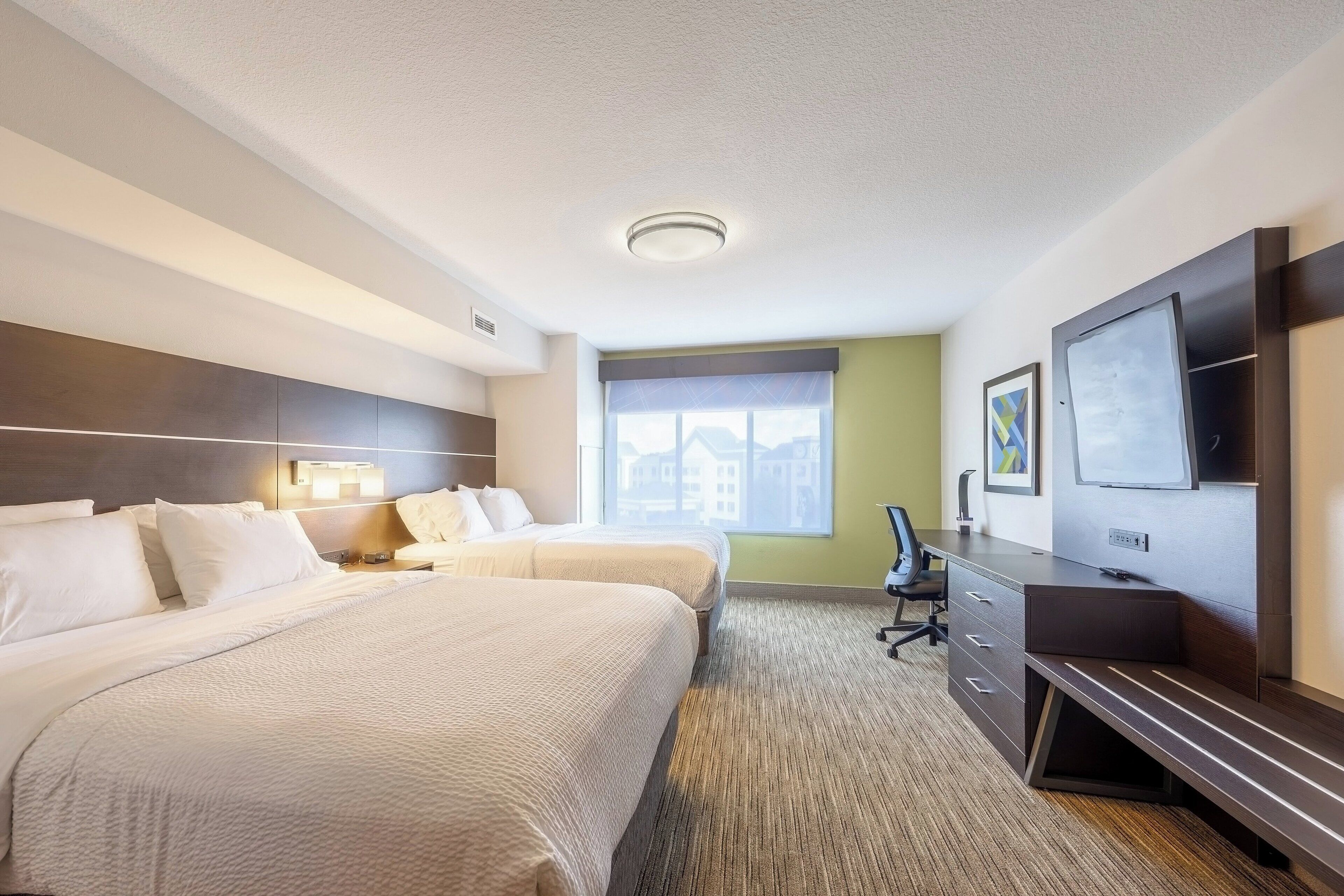 A hotel room with two beds, a desk, a chair, a flat-screen TV, and a window with a view of buildings.