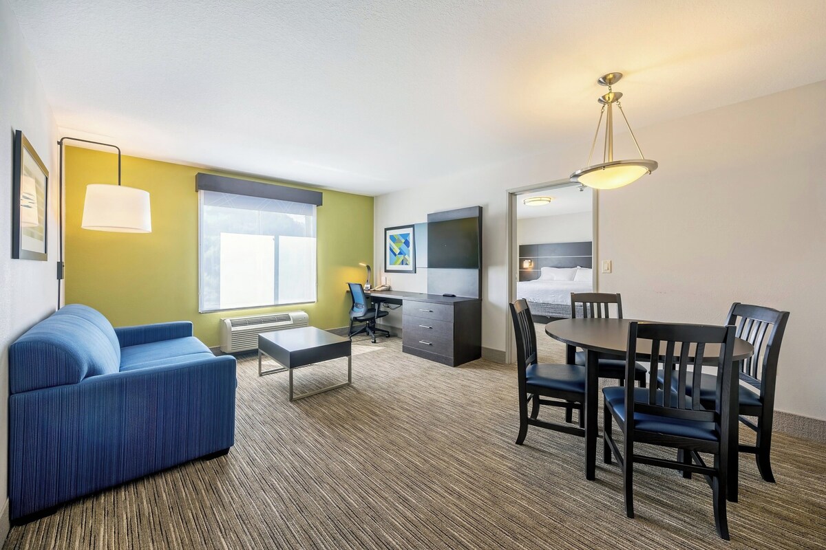 A hotel room with a blue sofa, a desk with a chair, a dining table with chairs, a TV, and a large mirror.