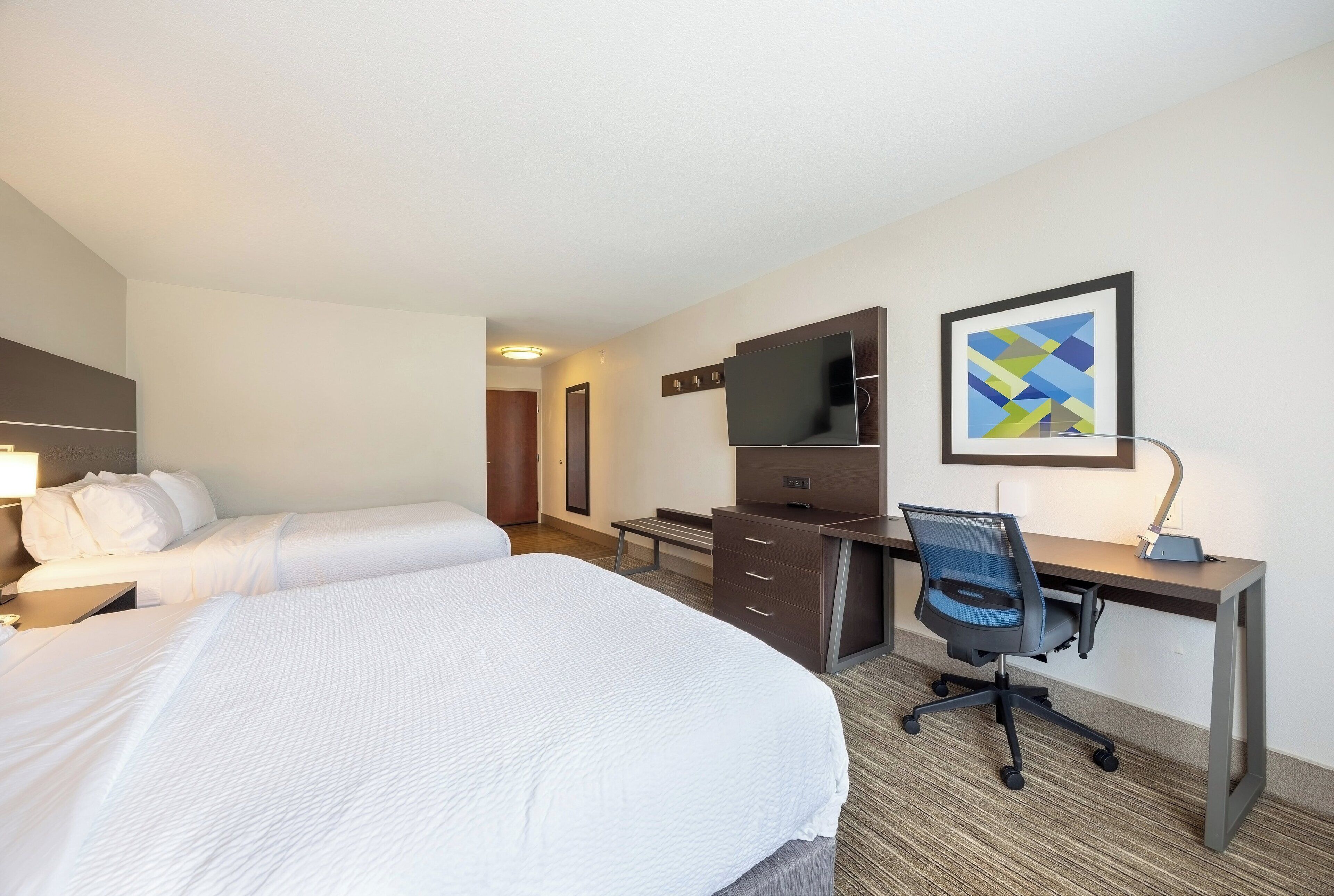 A hotel room with a large bed, a desk with a chair, a flat-screen TV, and a framed abstract art piece on the wall.