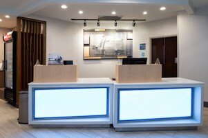 Zona de recepción de un hotel con dos mostradores, un amplio ventanal que muestra el nombre del hotel y un letrero que indica la ubicación del ascensor.