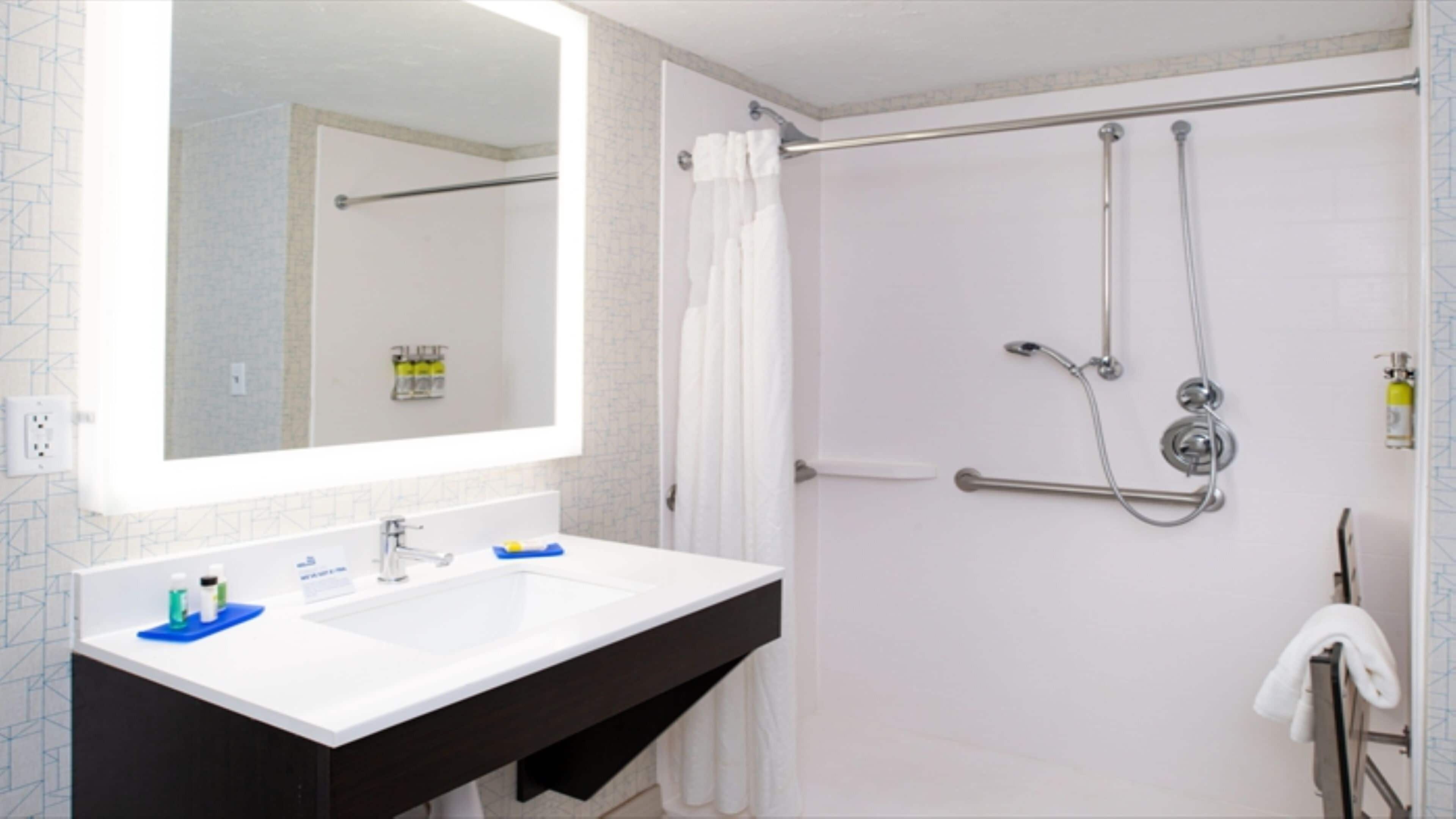 A bathroom with a white sink, dark countertop, and a large mirror. There is a shower with a handheld showerhead and a towel rack.