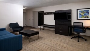 A modern hotel room with a blue sofa, a black office chair, a small black table, a TV on a stand, a desk with drawers, and a framed abstract art piece on the wall.