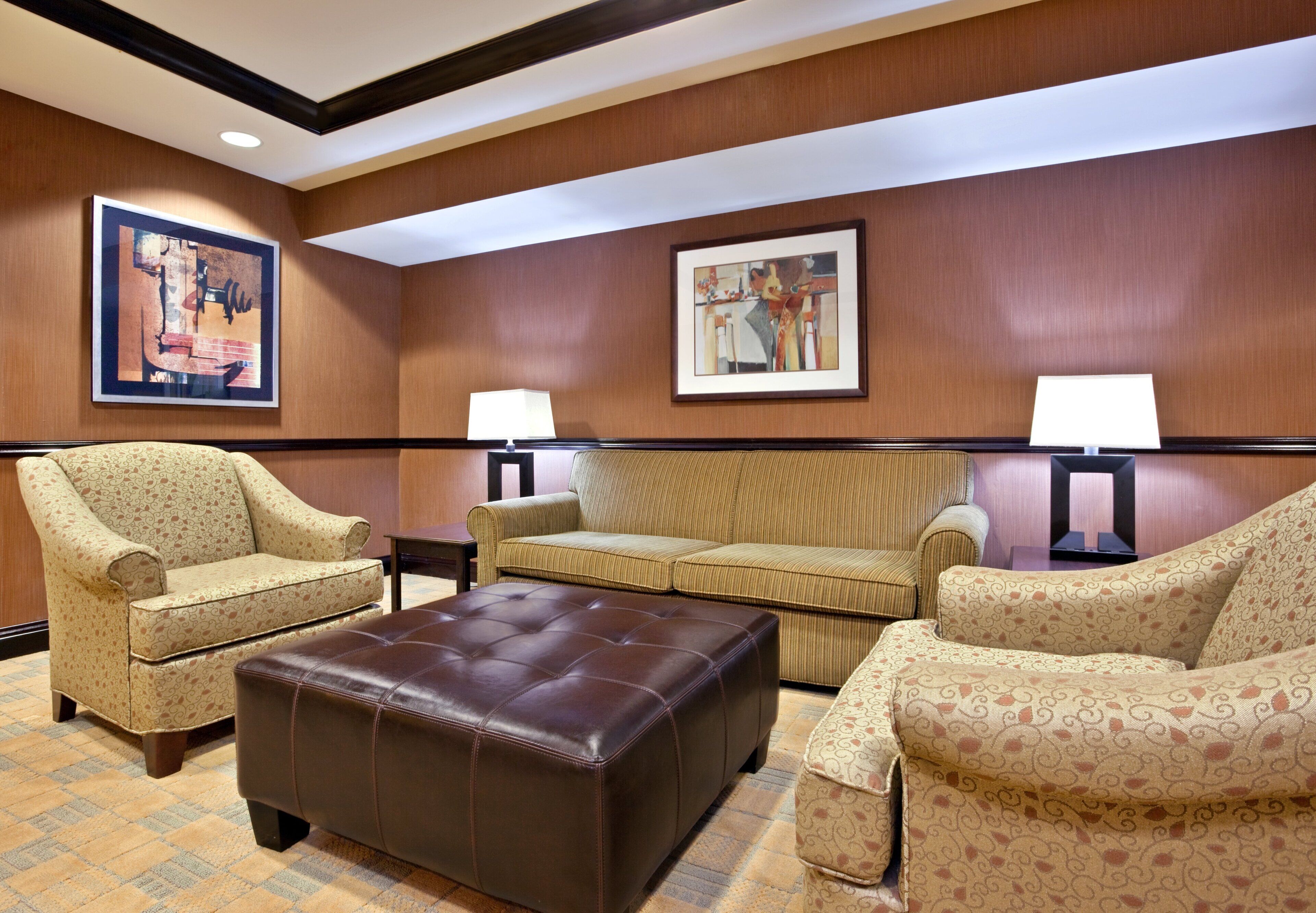 A hotel room with a brown leather ottoman, two patterned armchairs, a beige sofa, and two wall-mounted lamps.