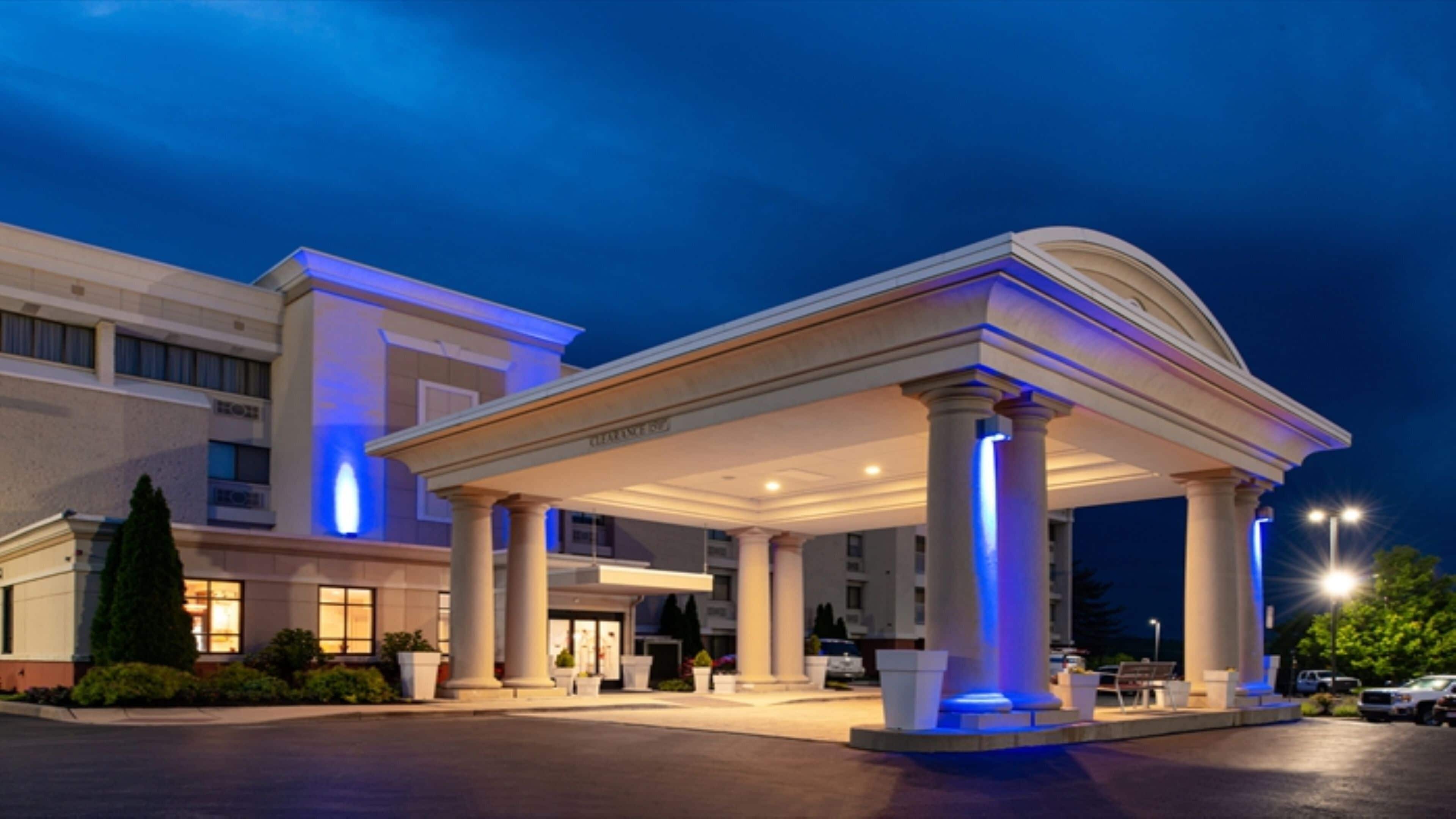 Entrada de hotel com área coberta sustentada por colunas, estacionamento e um edifício com janelas iluminadas.