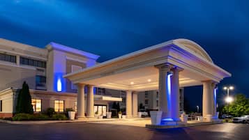 Entrada de hotel com área coberta sustentada por colunas, estacionamento e um edifício com janelas iluminadas.