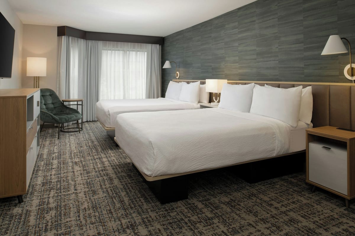 A hotel room with two beds, a desk, a chair, a TV, and a large window with curtains.