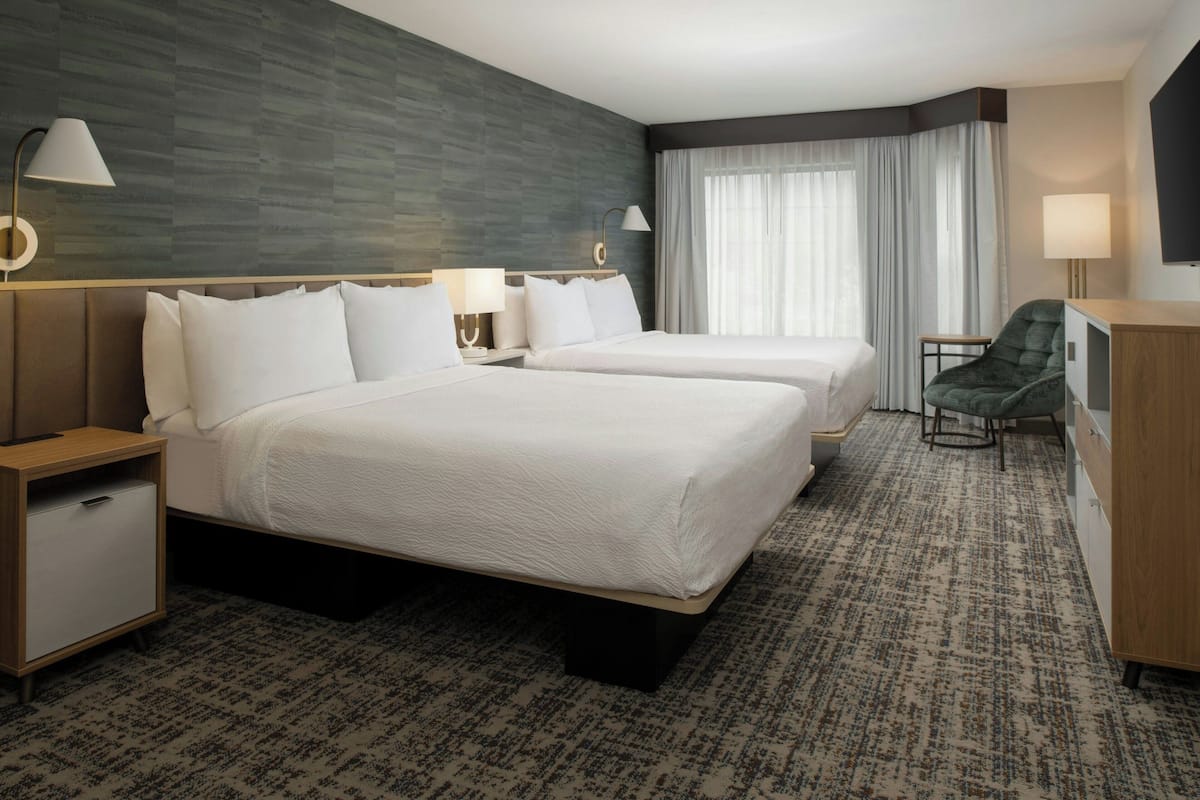 A hotel room with two beds, a desk, a chair, a TV, and a window with curtains.