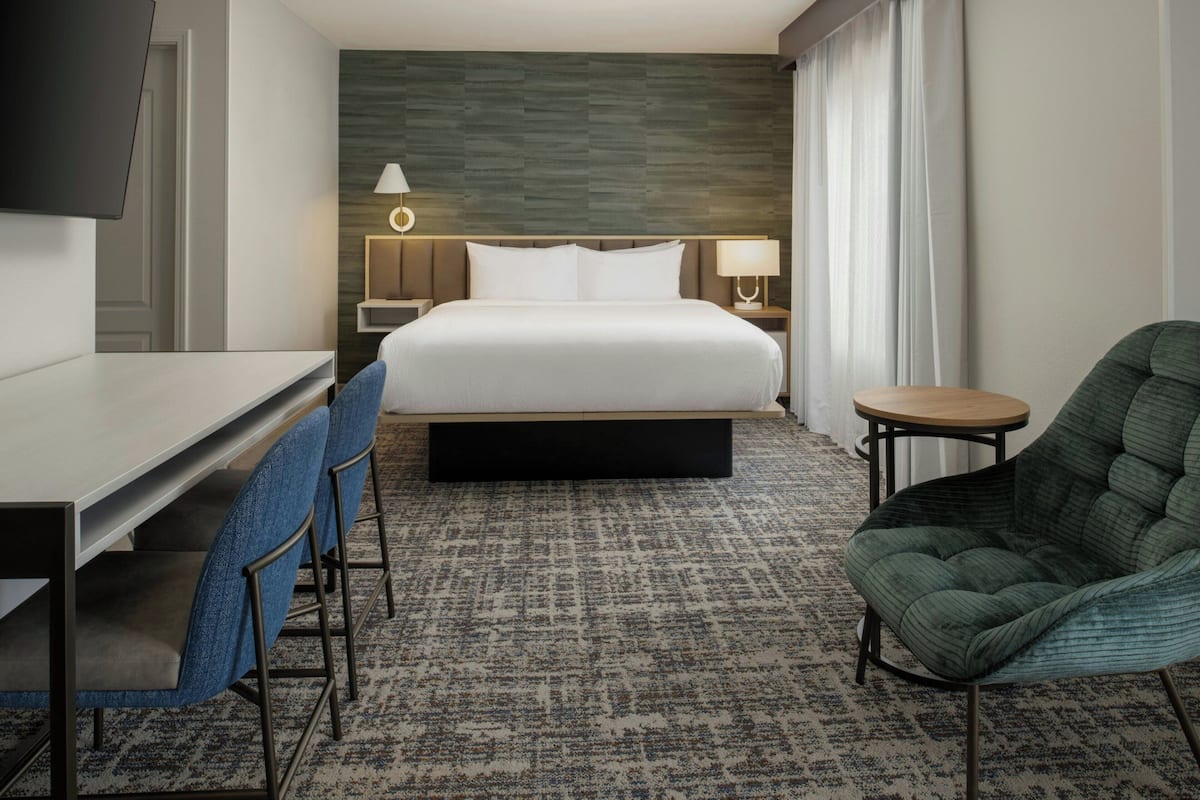 A hotel room with a bed, a desk, two chairs, a small table, and a TV mounted on the wall.