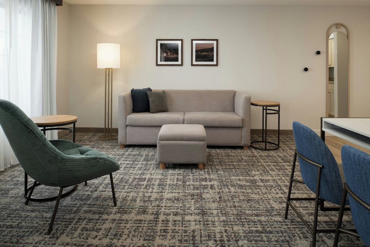 A hotel room with a sofa, ottoman, two chairs, a small table, a lamp, and two framed pictures on the wall.