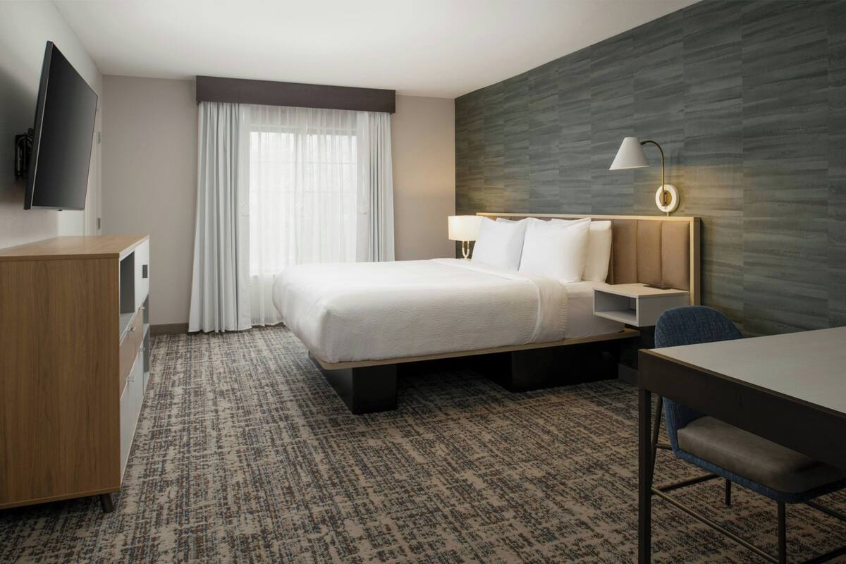 A hotel room with a bed, a desk, a chair, a TV, a lamp, and a window with curtains.