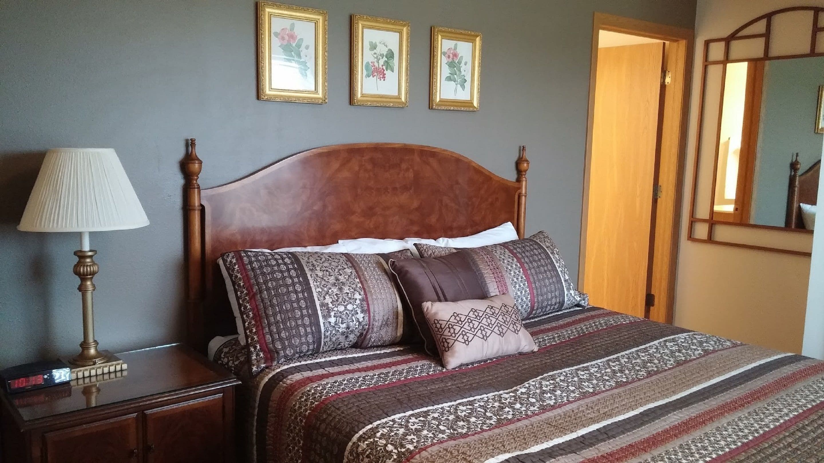Une chambre avec une tête de lit en bois, une table de chevet avec une lampe et une couvre-lit à motifs. Trois tableaux encadrés représentant des fleurs ornent le mur.