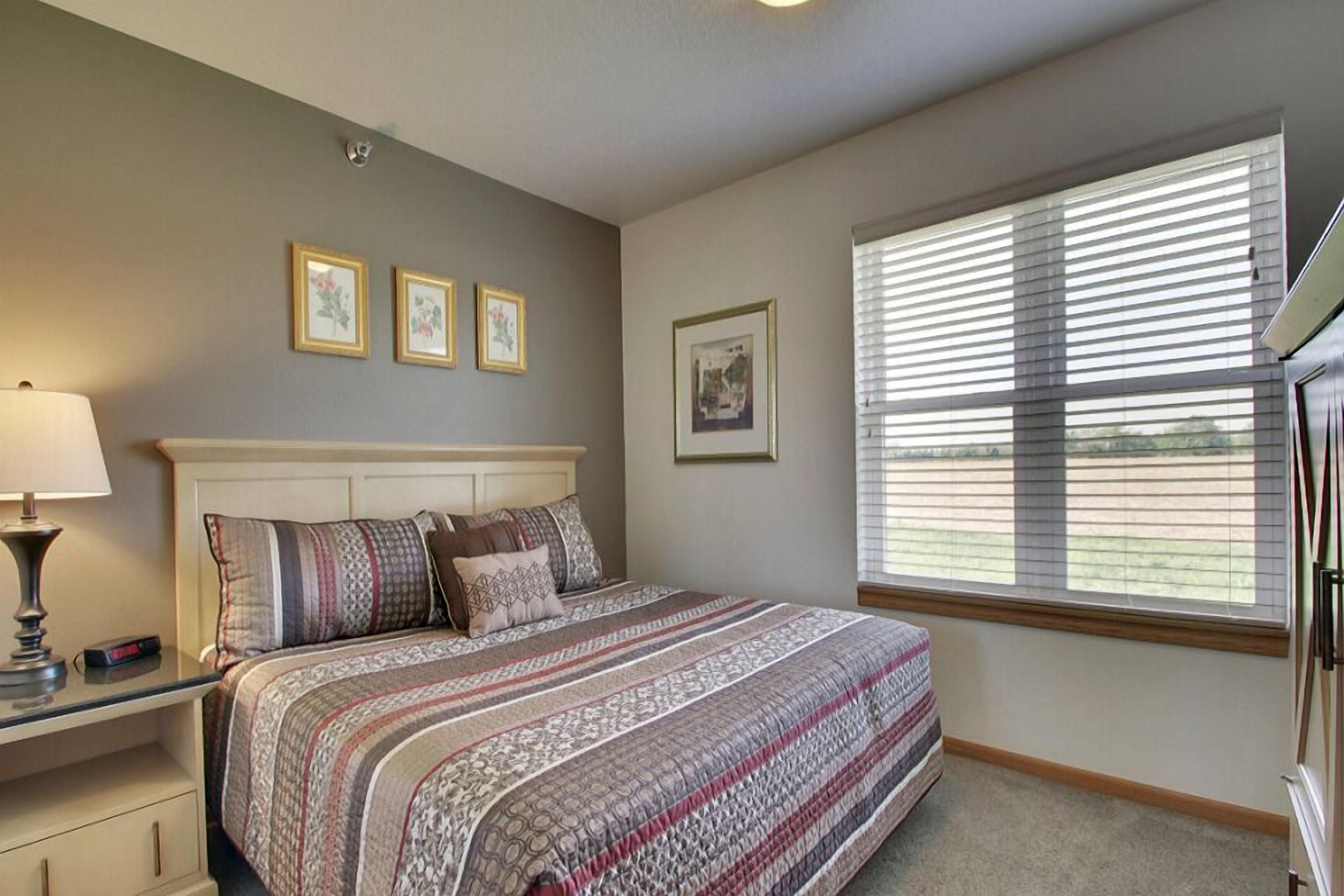 A bedroom with a bed, bedside table, lamp, and a window with blinds.