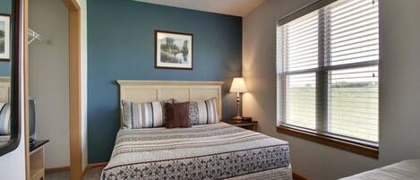 A bedroom with a large bed, a bedside lamp, a framed picture on the wall, and a window with blinds.
