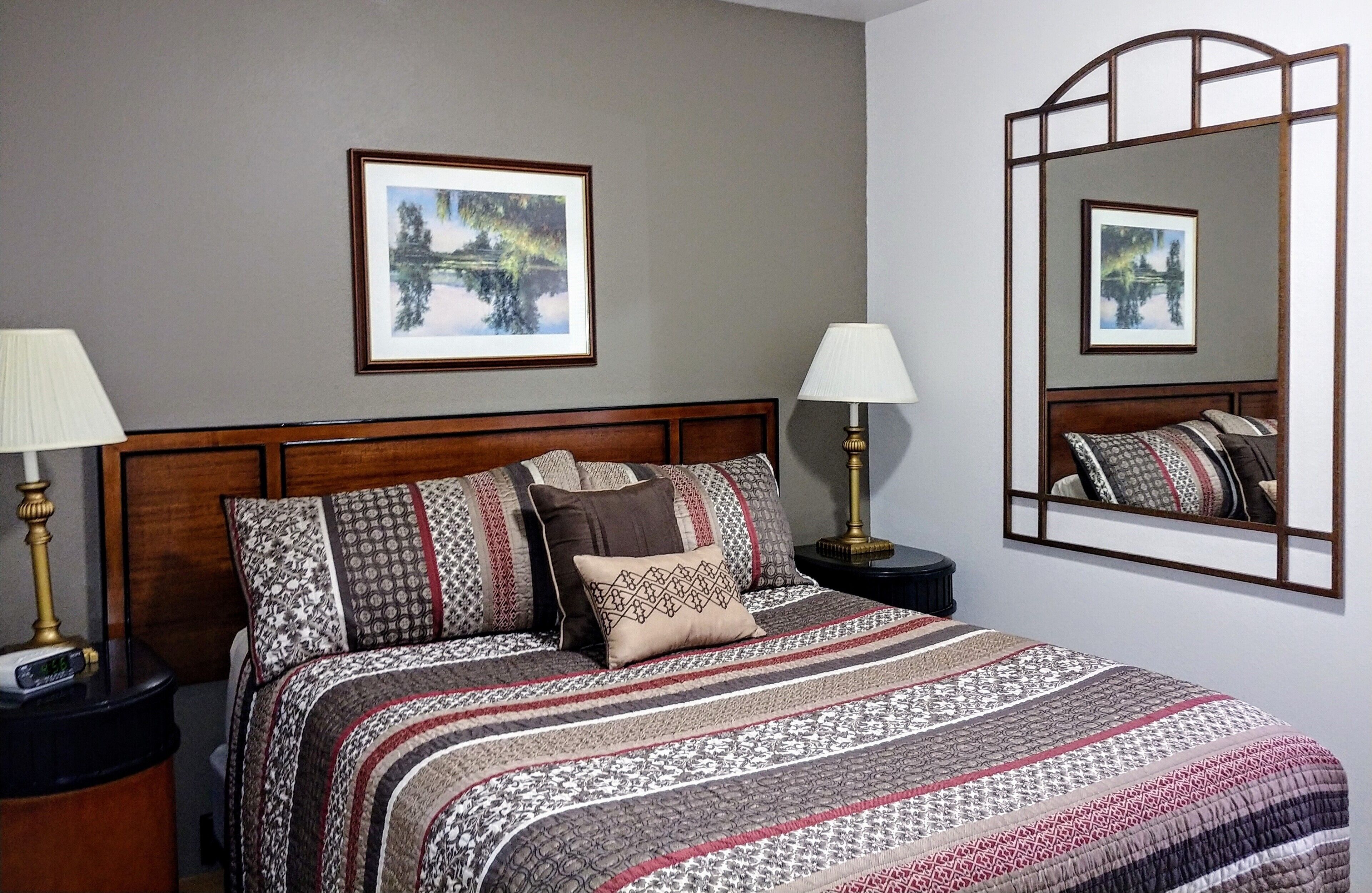 Uma cama bem arrumada com roupa de cama e almofadas estampadas, cabeceira de madeira, abajures e quadros na parede.