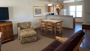 Ein Wohnzimmer mit einer Couch, einem gemusterten Sessel, einem Esstisch mit Stühlen, einer Küchenzeile mit Holzschränken und einem Kühlschrank sowie einem Fernseher auf einem Standfuß.