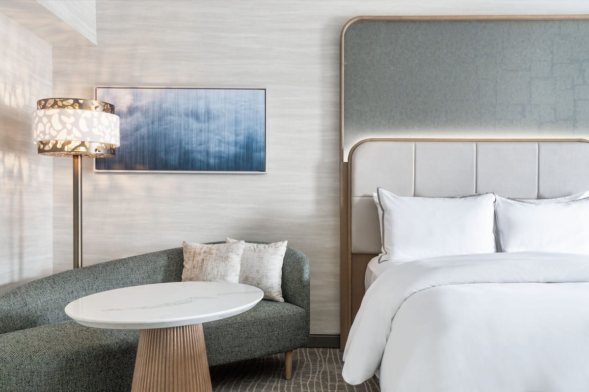 A modern hotel room with a grey upholstered sofa, a round white table, a large bed with white linens, and a contemporary lamp.