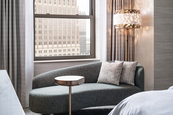 Una habitación de hotel moderna con un sofá gris curvo, una mesita redonda pequeña y un ventanal con vistas a un paisaje urbano.