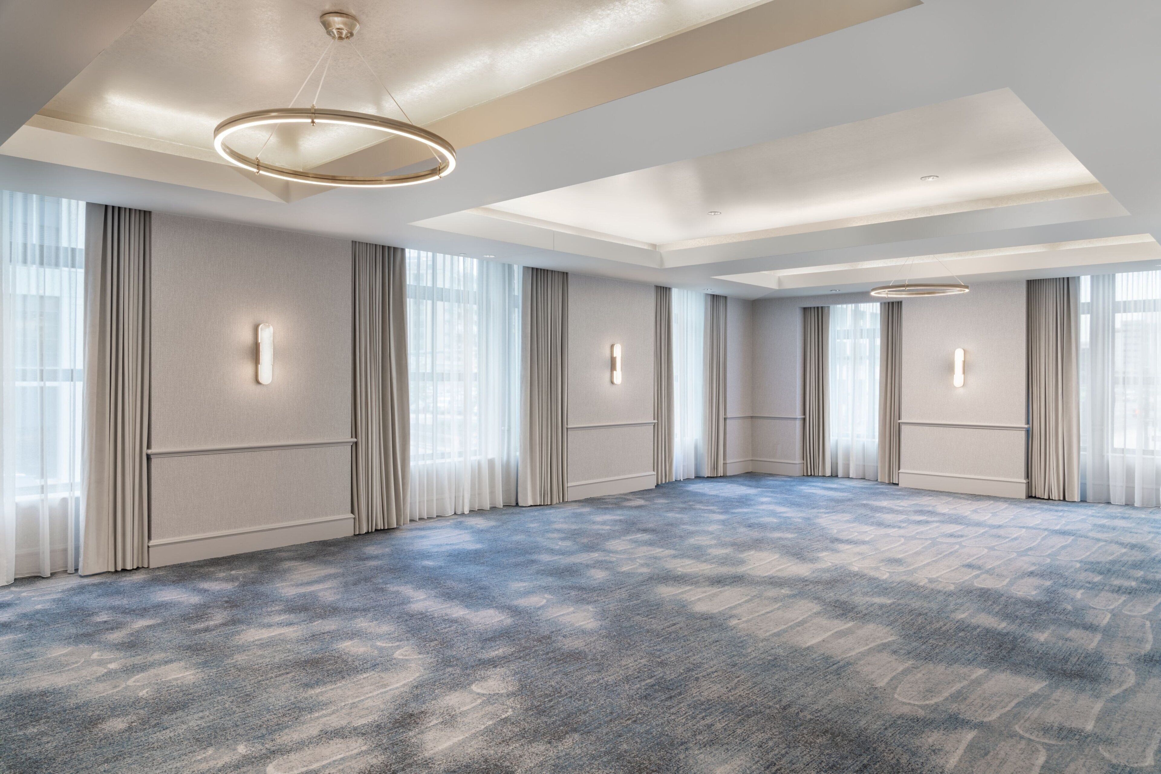 A spacious hotel room with a large carpeted area, large windows with curtains, and a modern chandelier.