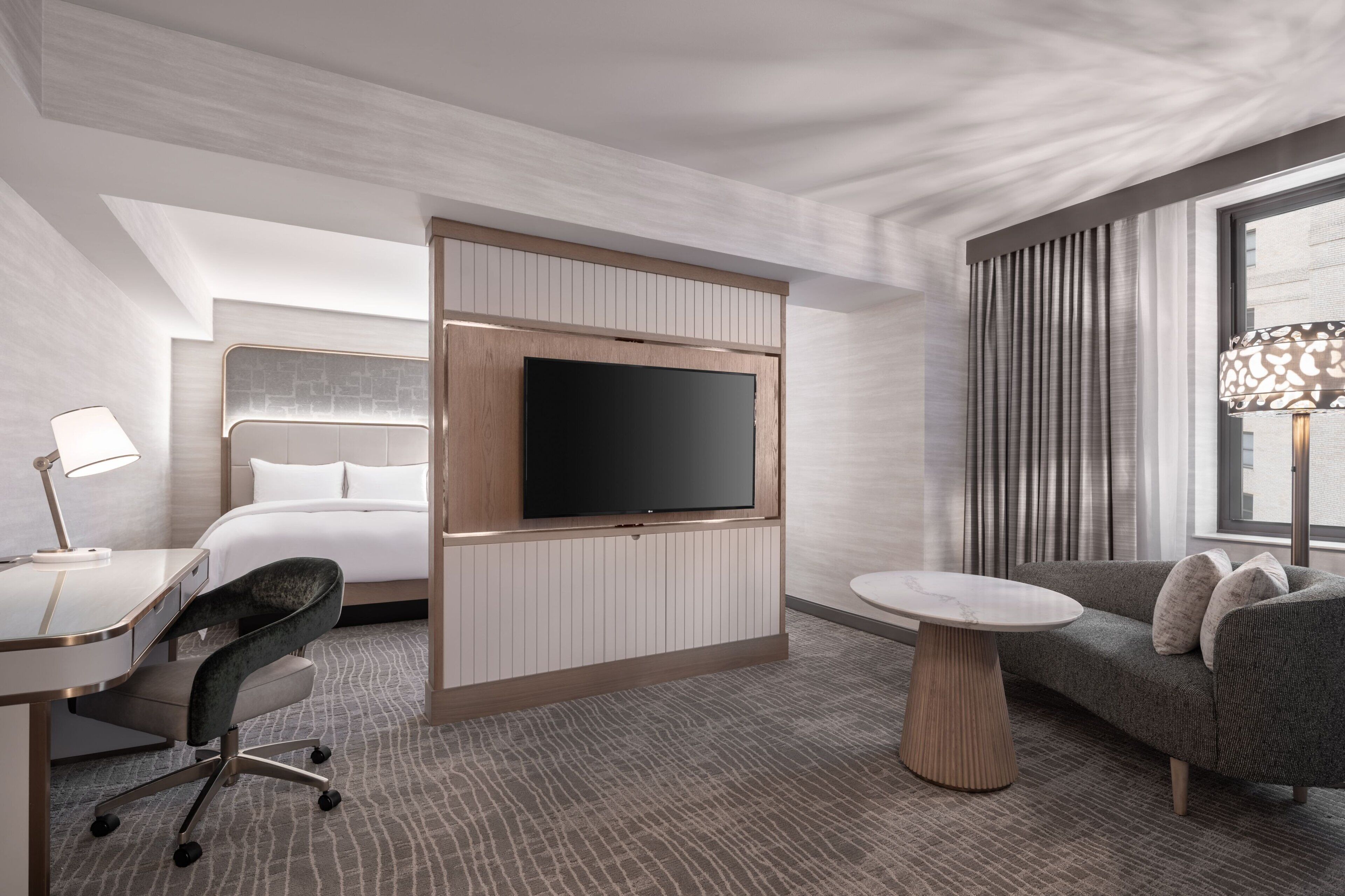 A modern hotel room with a bed, a TV mounted on a wooden panel, a desk with a chair, a sofa, and a lamp.