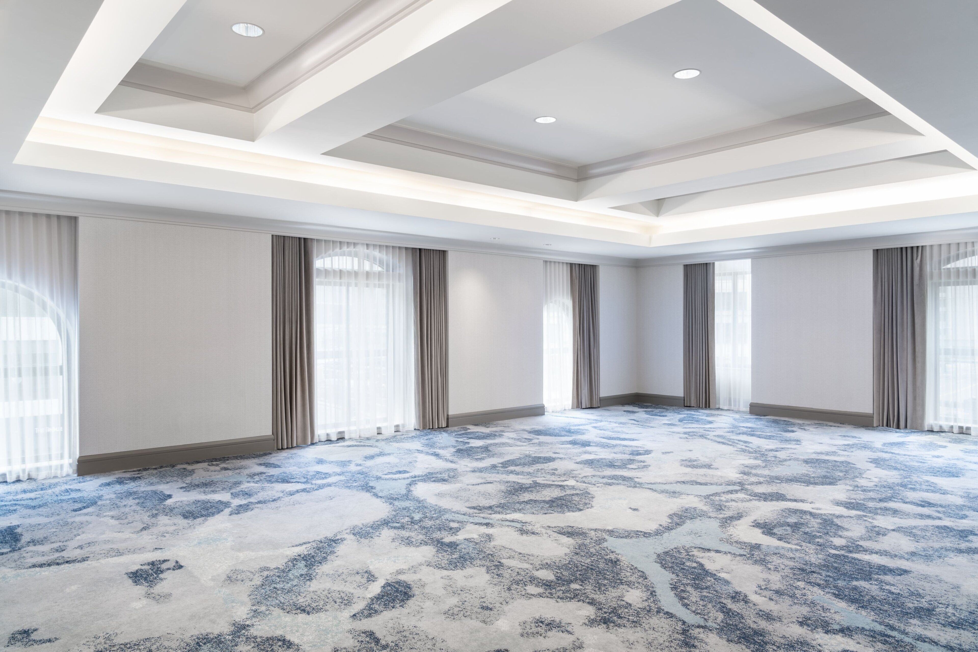 A spacious hotel room with a large carpeted area, a high ceiling with recessed lighting, and large windows with curtains.