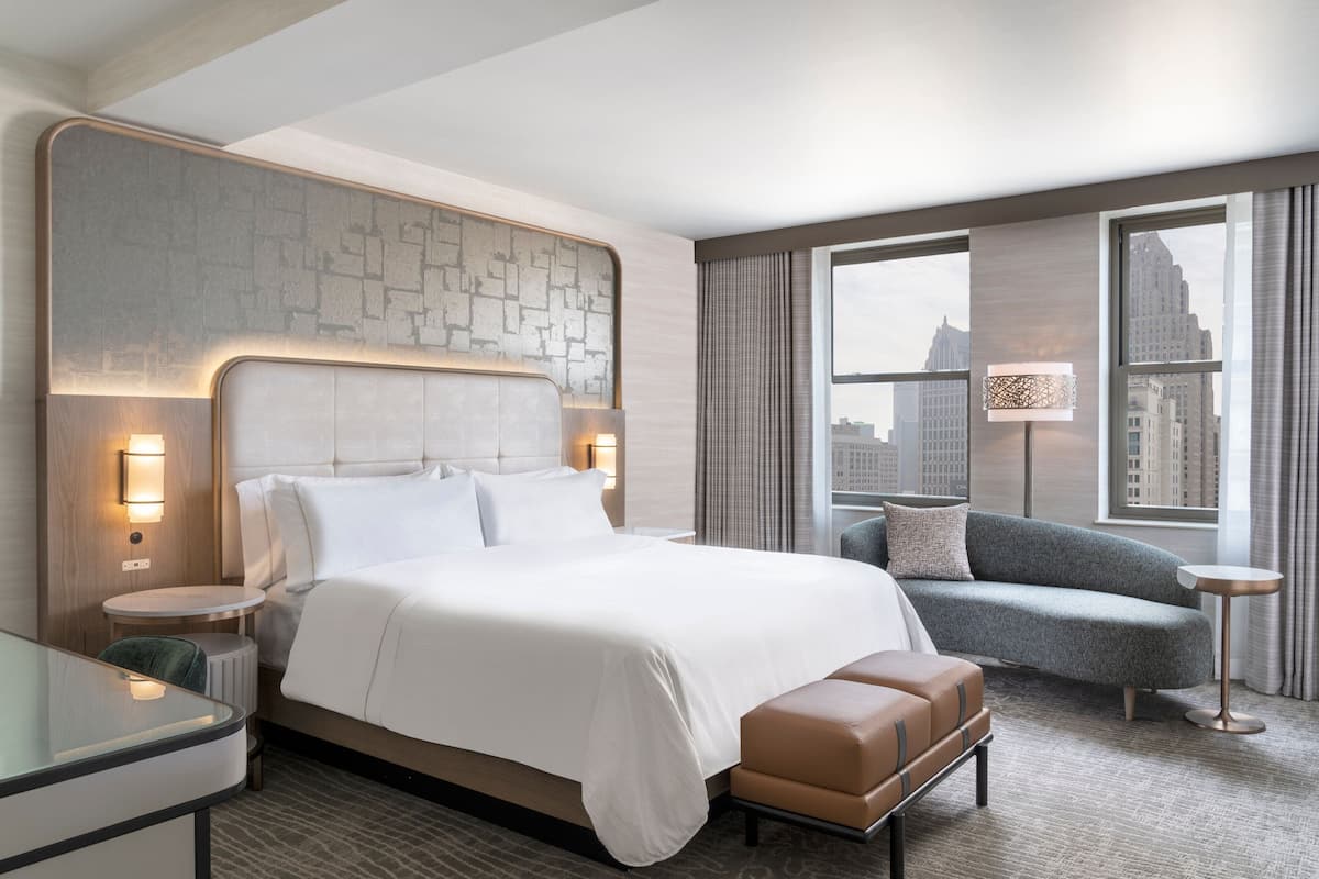 A modern hotel room with a large bed, a sofa, a small table, and a view of the city.