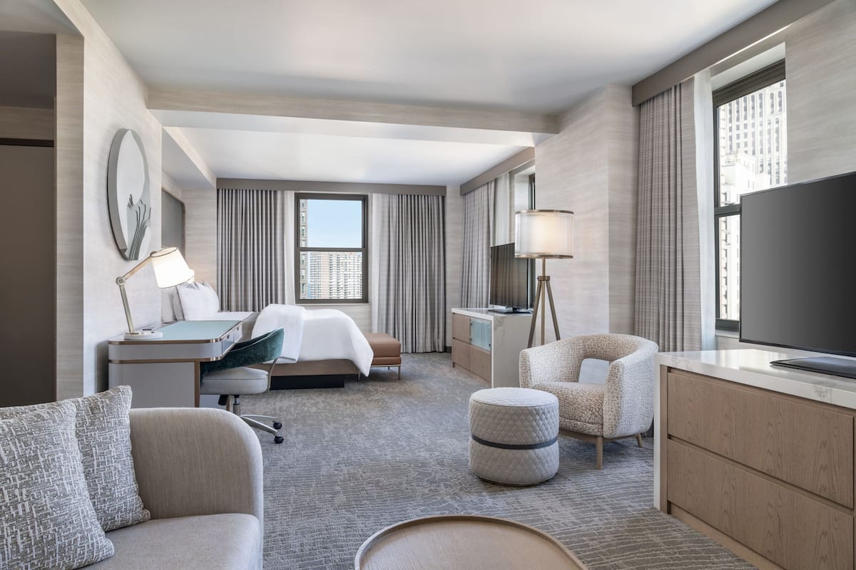 A hotel room with a bed, desk, chair, ottoman, TV, and large windows.