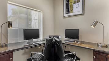 A home office with two desks, each with a computer and a lamp. There are two chairs, one with a backrest and one without. A framed abstract painting is on the wall.