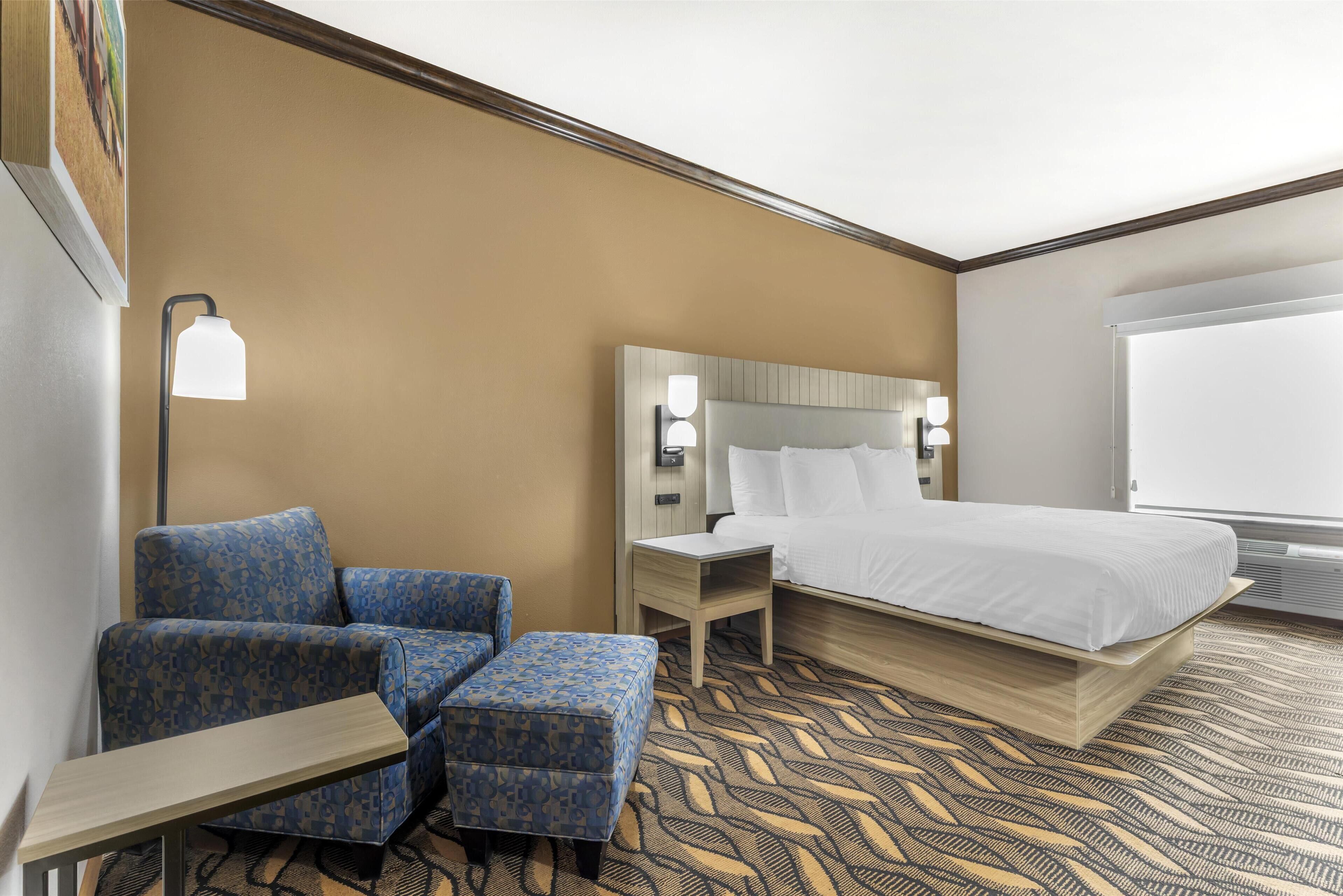 A hotel room with a bed, a blue patterned armchair, a small table, and a nightstand with a lamp.