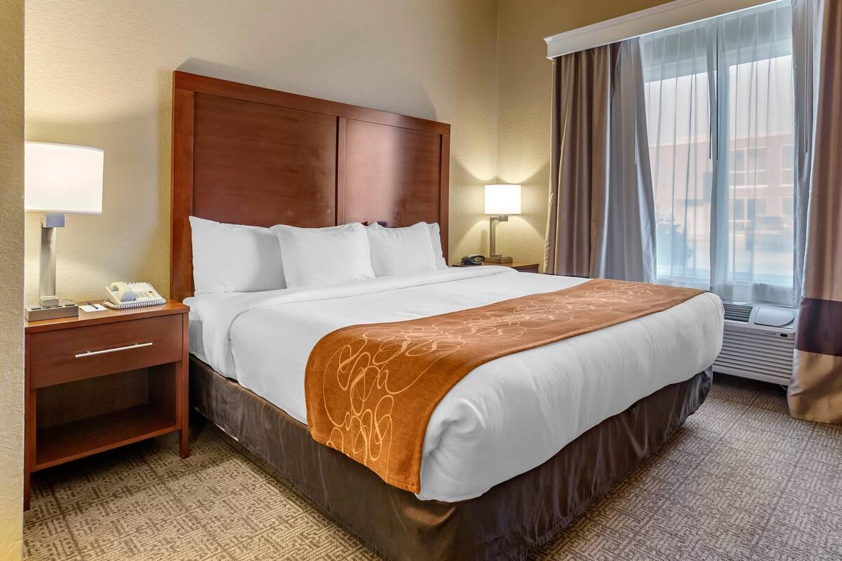 A hotel room with a large bed, a bedside table with a lamp, a window with curtains, and an air conditioning unit.
