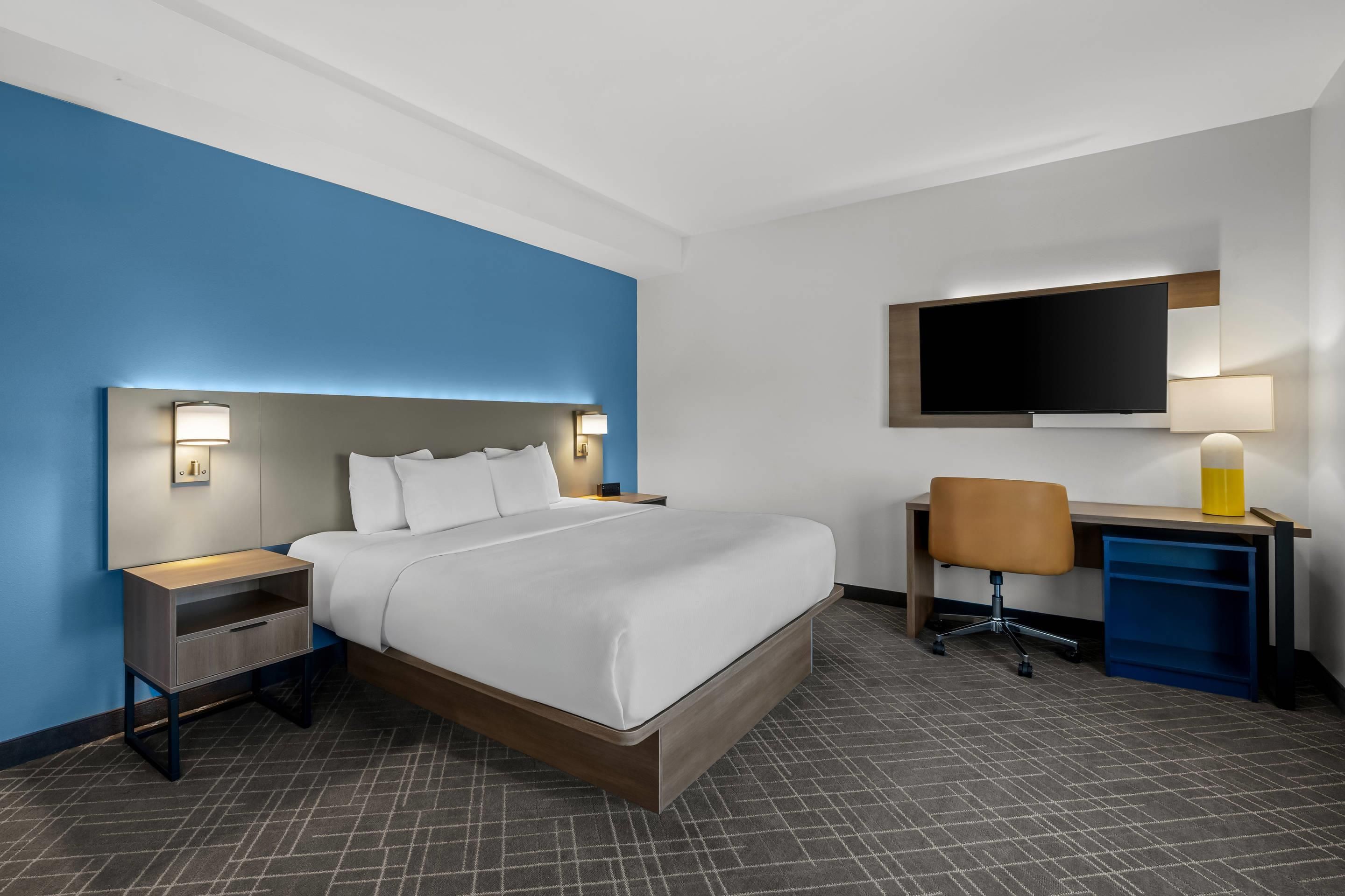 Quarto de hotel com cama, mesas de cabeceira, escrivaninha com cadeira, televisão de tela plana e abajur.