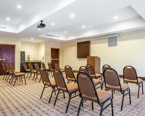 Een congreszaal met vloerbedekking, rijen stoelen, een kitchenette en een plafondprojector.