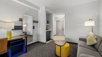 Een hotelkamer met een bank, een klein tafeltje, een spiegel, een lamp, een prullenbak en een deur naar een badkamer.