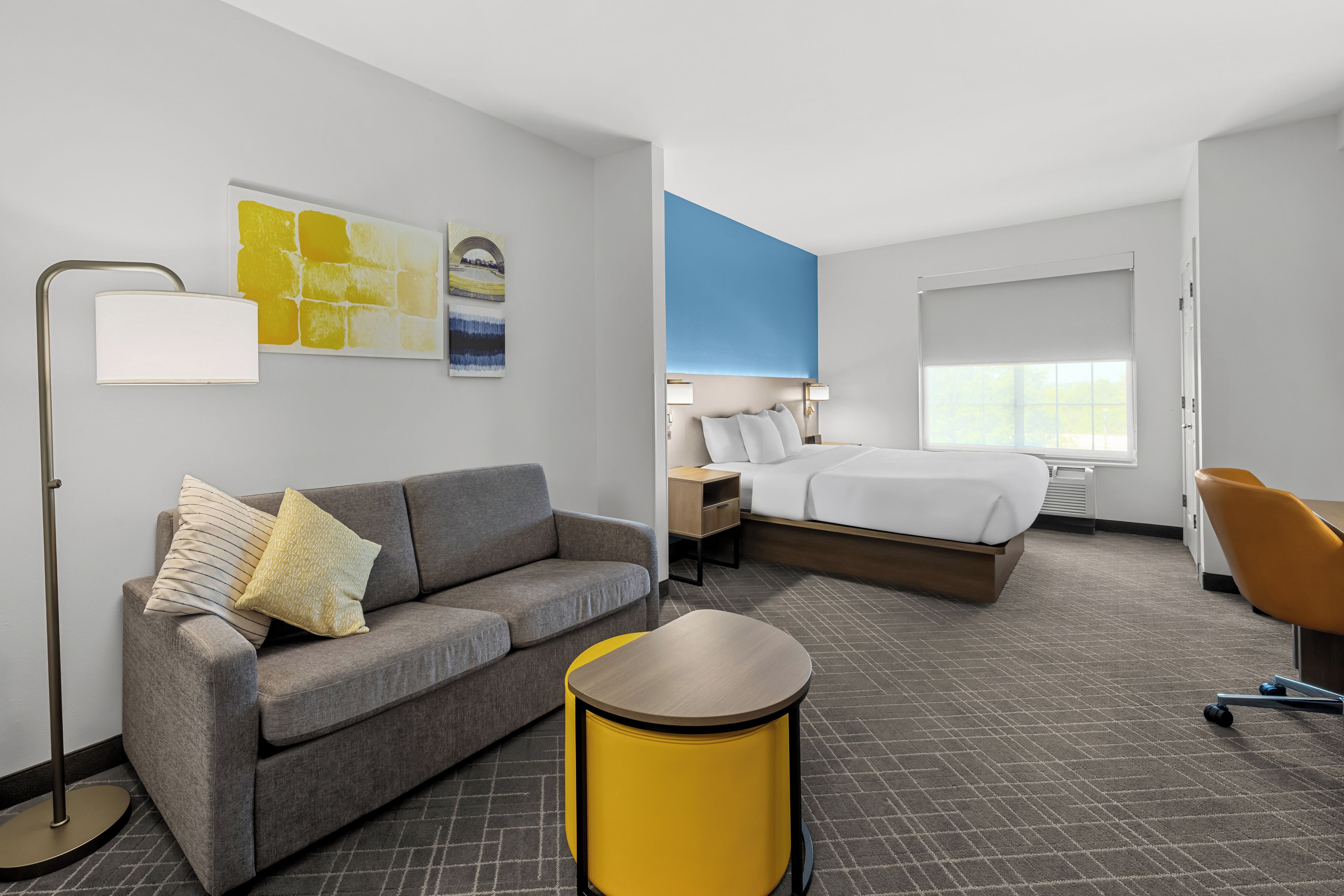 Een hotelkamer met een grijze bank, een geel bijzettafeltje, een bed met wit bedlinnen, een blauwe accentmuur en een groot raam.