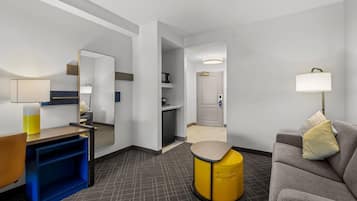 A hotel room with a desk, chair, sofa, and a small yellow stool.