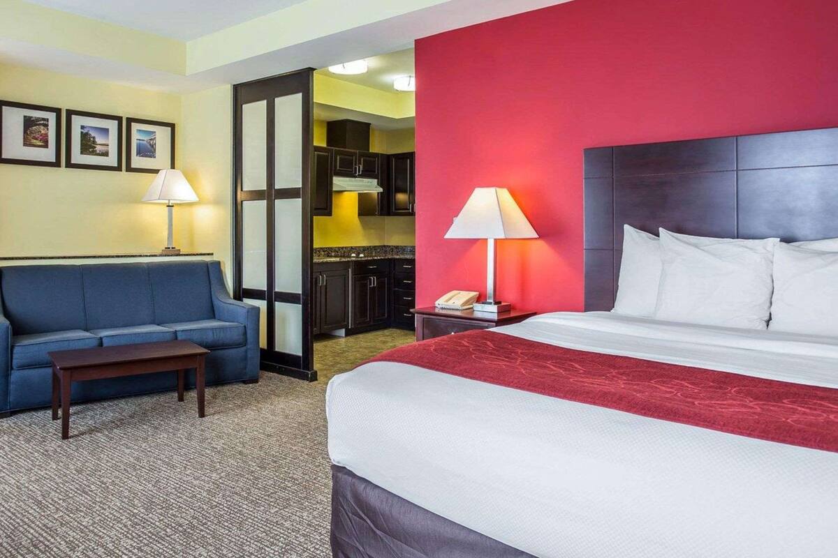 Une chambre d’hôtel avec un lit, un canapé bleu, une petite table, une table de chevet avec une lampe et une vue sur la cuisine.