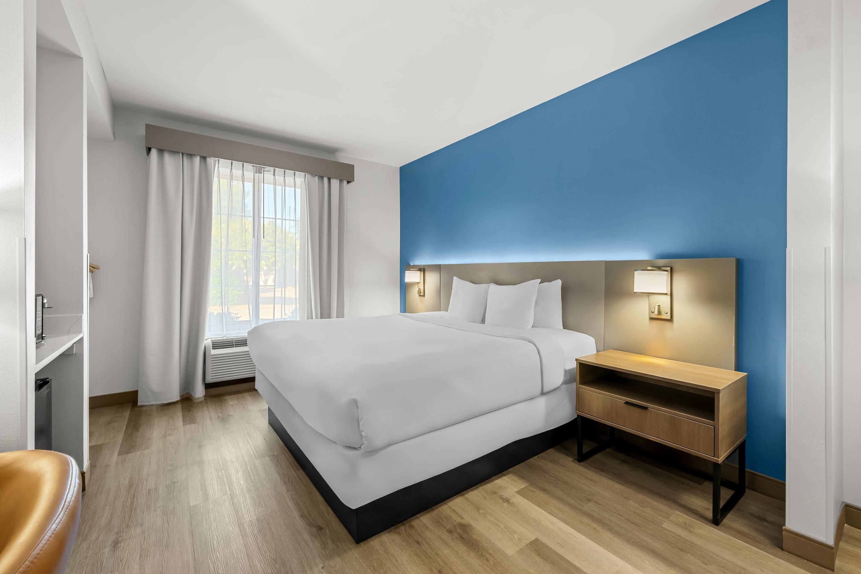 Habitación de hotel con una cama grande, una mesita de noche con lámpara, un armario y una ventana con cortinas.
