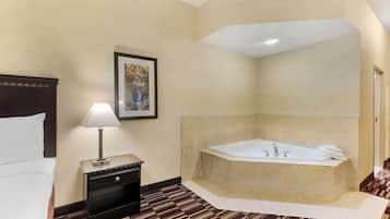 A hotel room with a large bed, a bedside table with a lamp, a framed picture on the wall, a large bathtub, and a patterned floor.