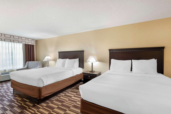 A hotel room with two beds, a chair, a nightstand with a lamp, and a window with curtains. - Best Western Plus Victor Inn & Suites (Victor)