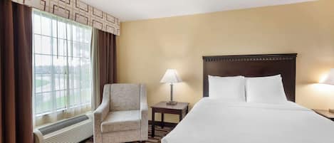 Habitación de hotel con cama, una silla, mesita de noche con lámpara, ventana con cortinas y un calentador en la pared.