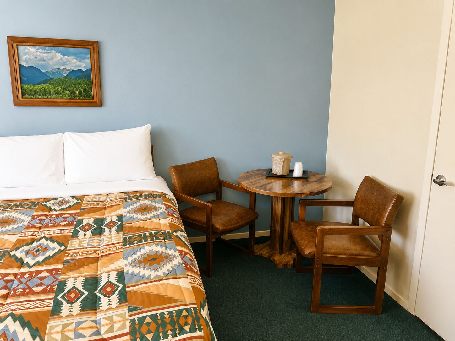 A hotel room with a bed, two chairs, a small table with a teapot, and a framed picture on the wall.