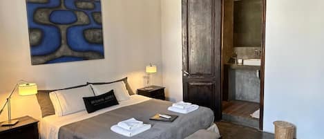 Un dormitorio con una cama grande, mesitas de noche, un banco, una obra de arte en la pared y una puerta que da a un baño.