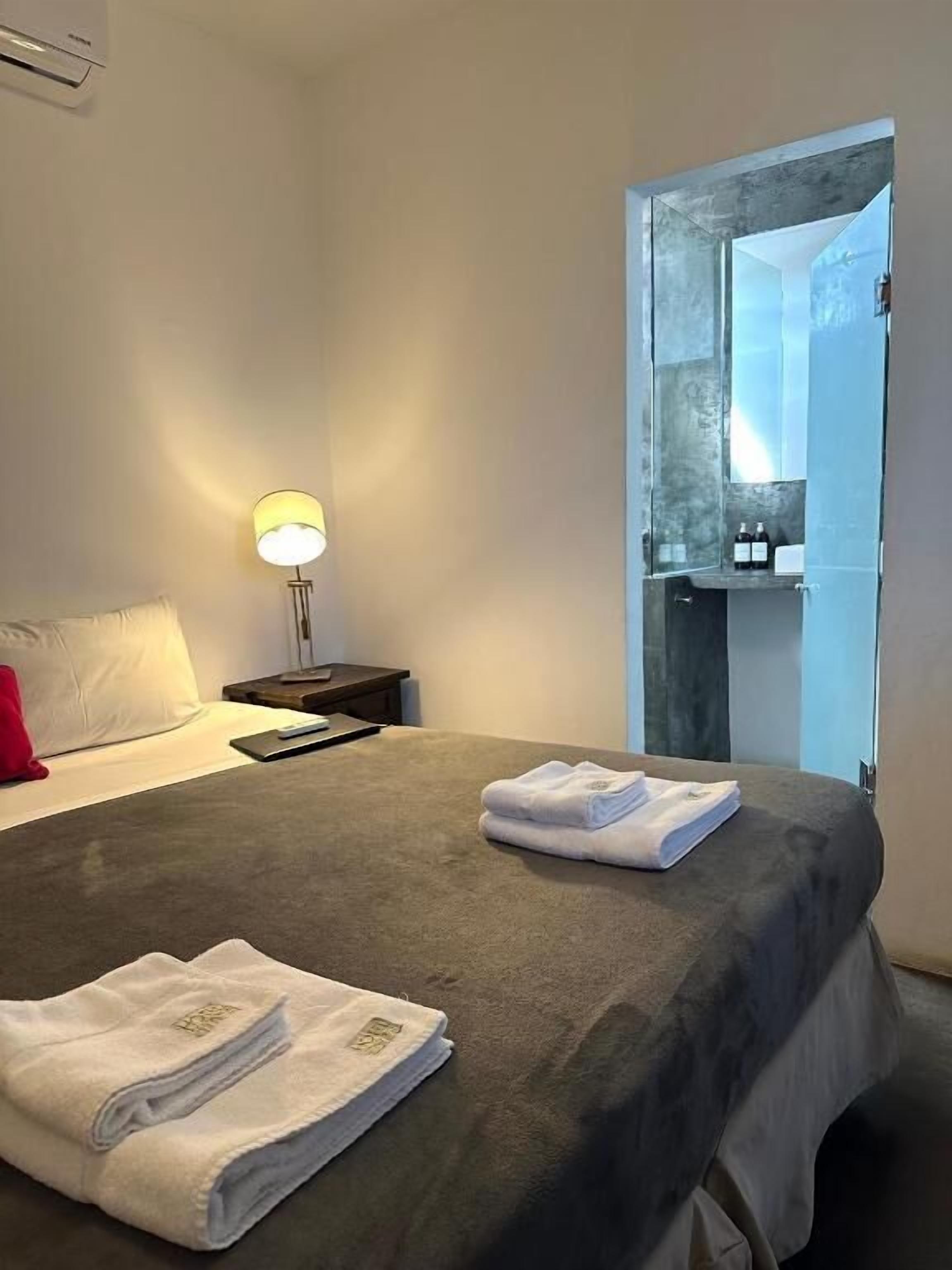 A hotel room with a bed, a nightstand, a lamp, and a bathroom visible through a glass door.