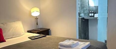 A hotel room with a bed, a nightstand, a lamp, and a bathroom visible through a glass door.