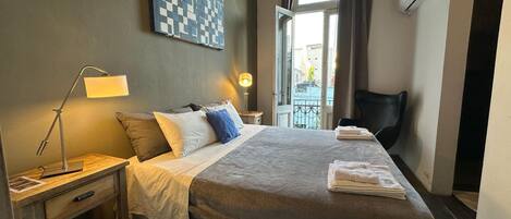 Una cama bien hecha con una manta, almohadas y una lámpara de noche. Hay una mesita con una lámpara y un control remoto. Se ve una ventana con cortinas y un equipo de aire acondicionado.