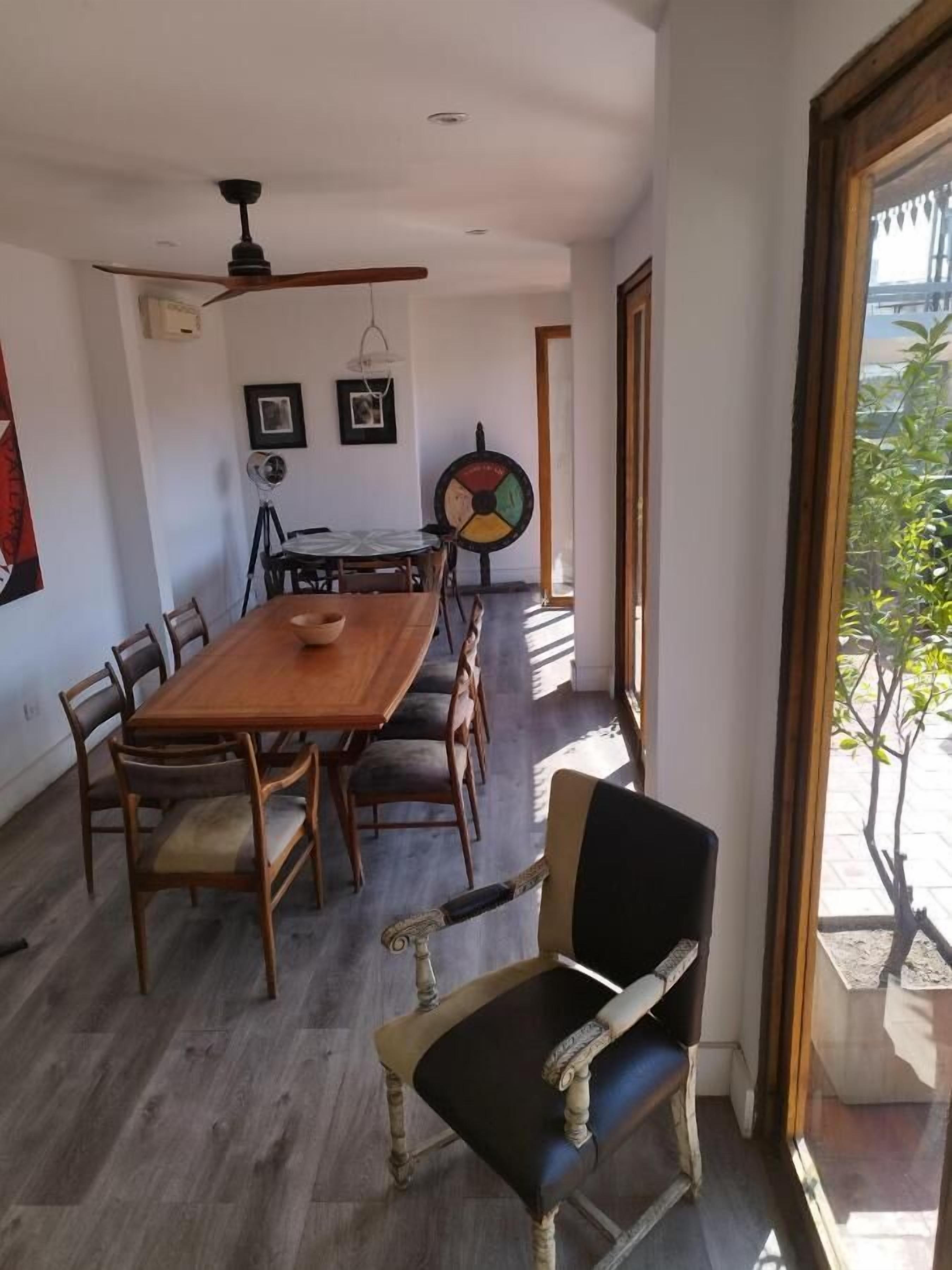 Zona pranzo con tavolo e sedie in legno, ventilatore a soffitto e finestra vista verde.
