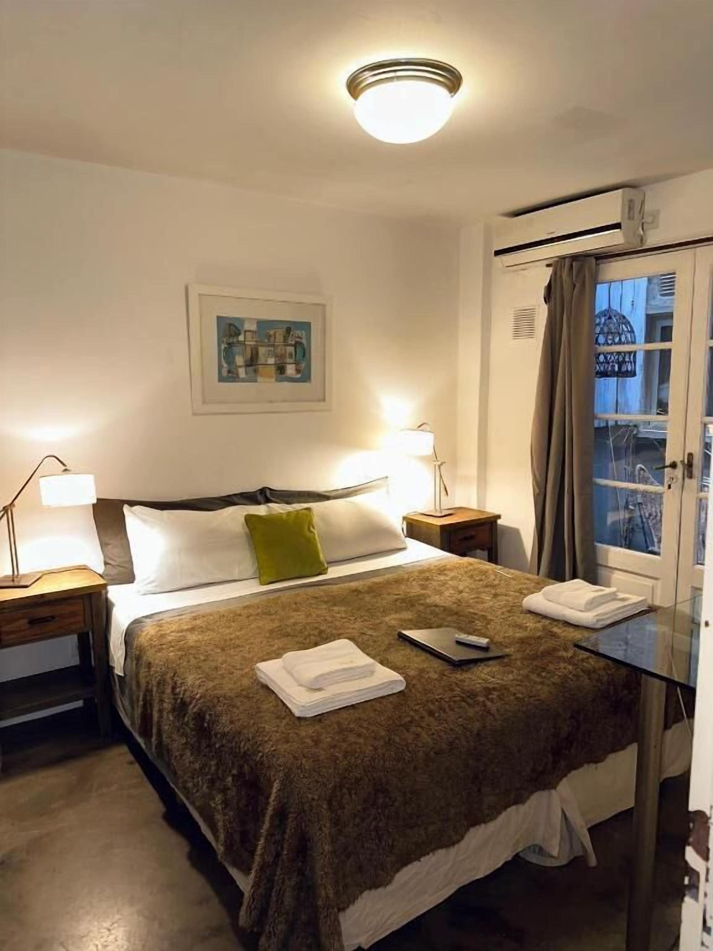 Een hotelkamer met een bed, twee bedlampen, een raam met gordijnen, een wandairconditioner en een ingelijst kunstwerk aan de muur.