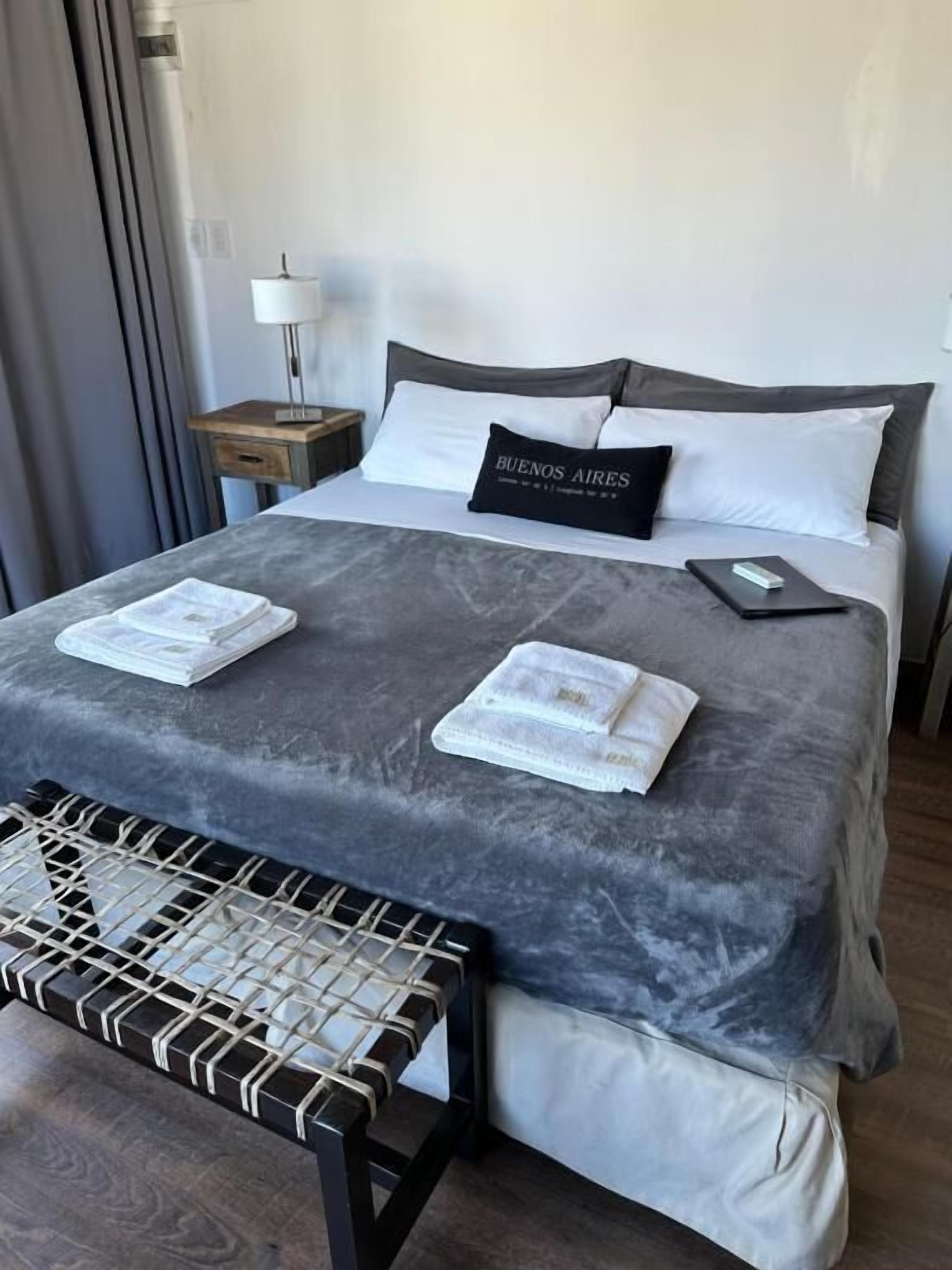 Een net opgemaakt bed met een grijze spreidek, twee kussens, en een zwart kussen met tekst. Er staat een klein nachtkastje met een lamp, een zwart dienblad met een zakdoekendoos, en een zwart-wit gestreept bankje.