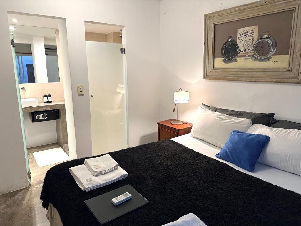 Habitación de hotel con cama, mesita de noche, lámpara, un cuadro en la pared y un baño visible a través de una puerta abierta.