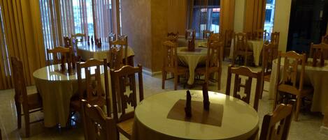 A dining area with multiple round tables and wooden chairs, featuring chandeliers and large windows with curtains.